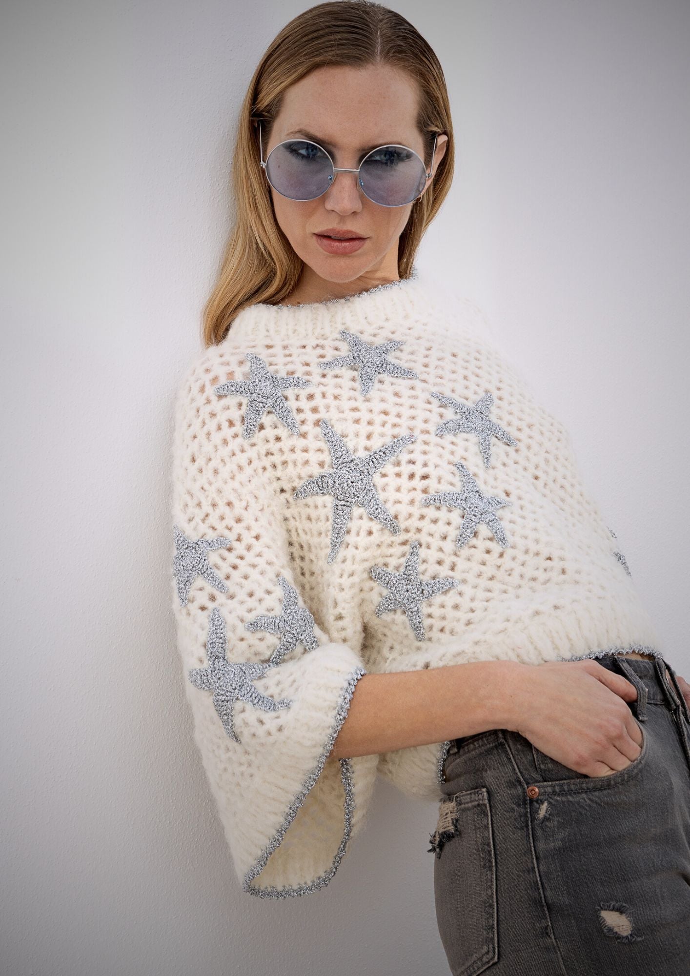 Girl leaning agianst a wall wearing a crocheted creme top, with vintage jeans